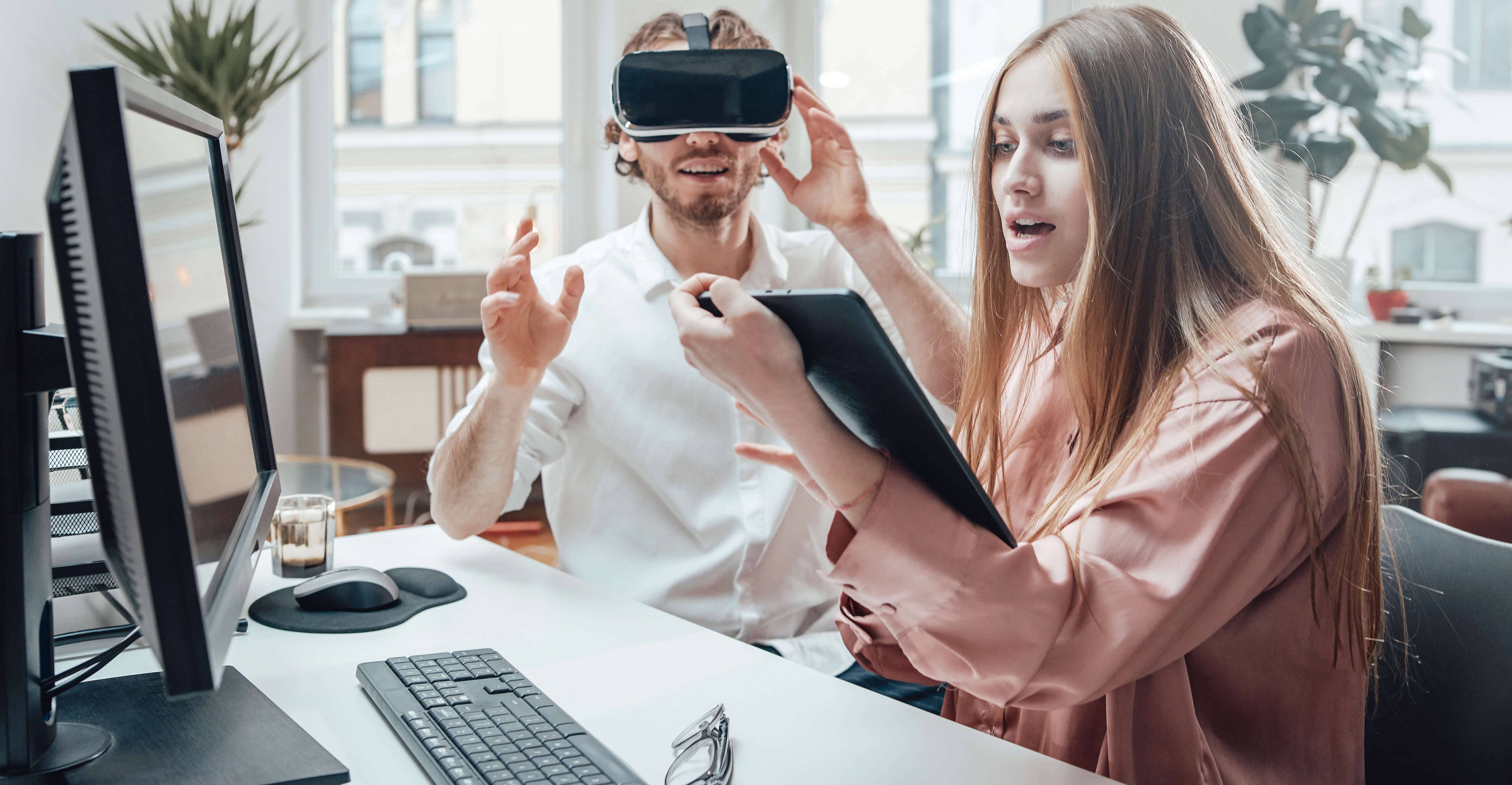 Guy dressed with virtual reality glasses is having fun at work with his female colleague who is using the tablet in the coworking office