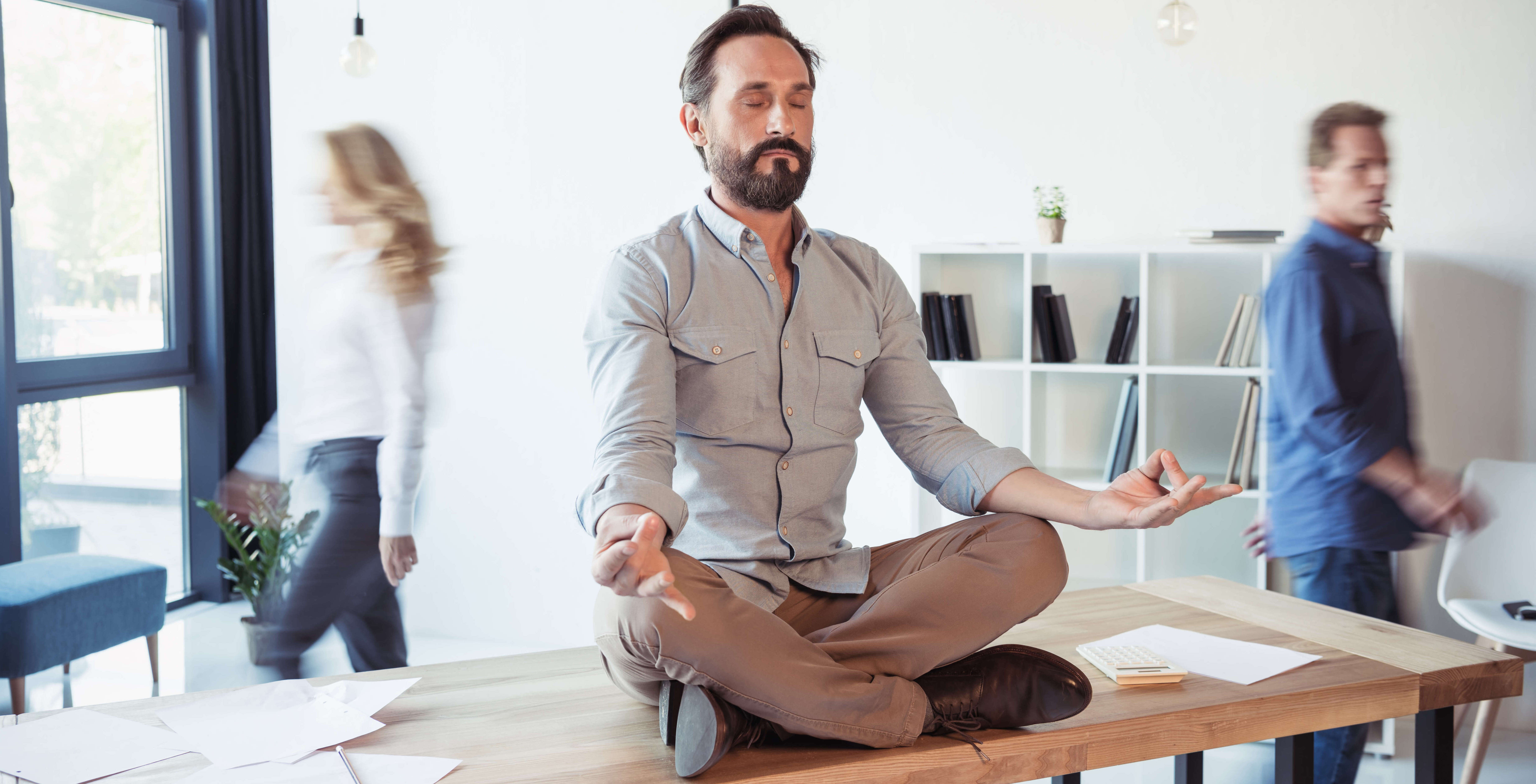 ​Homme d'affaires méditant en position de lotus pendant que ses collègues se déplacent dans le bureau de coworking